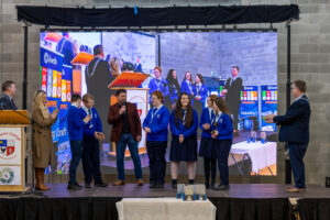 Six Students from Dunamase College in blur uniforms being interviewed on stage with a live strema of the event playing on the big screen behind them.