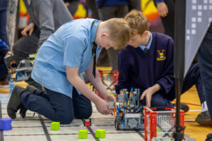 Two secondary school students in blue uniforms kneeling on the Vex Robotiics competition arena as they they work on their Vex robot