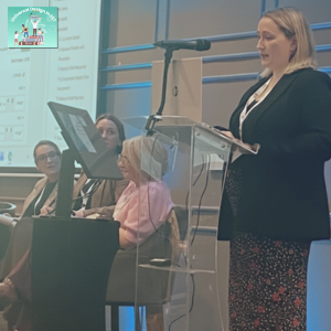 ETBI UD in FET Event Photo: This photo has 4 members of LOETB staff. Anne Marie Keane (QA FET Manager) is presenting at the podium. Sitting next to her is Thérèse Glennon (LOETB FET Access Officer), then Mary Lanier (LOETB FET Teacher) and on the far left is Jolene Hall (Enterprise Skills FET Manager). There is a large presentation screen next to Jolene Hall. In the top left corner there is a logo with the words Universal Design in FET, and it has an animated graphic of students standing on top of stacked books and holding other books.