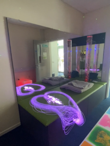 Sensory room A:  This is a photo of a sensory room in LOETB, there is wall mounted mirrors on two walls behind a padded green plinth.  On the plinth is a grey weighted blanket and strands of fibre optic purple lights, designed in the shape of a heart. 